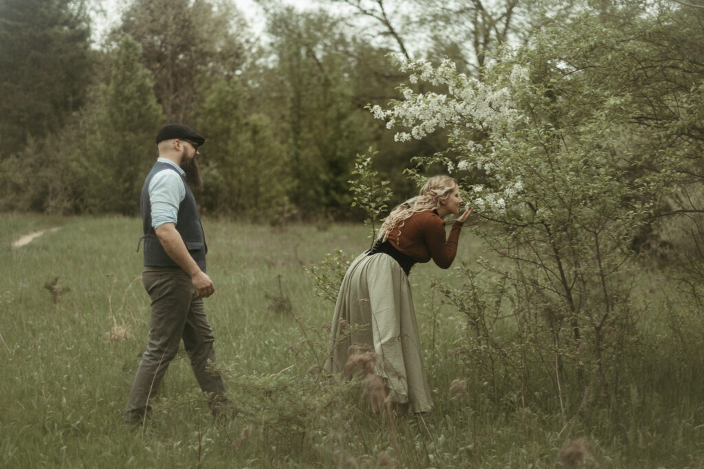 A woman skipping as she goes to a flower tree with her husband walking behind her at her engagement session in Michigan