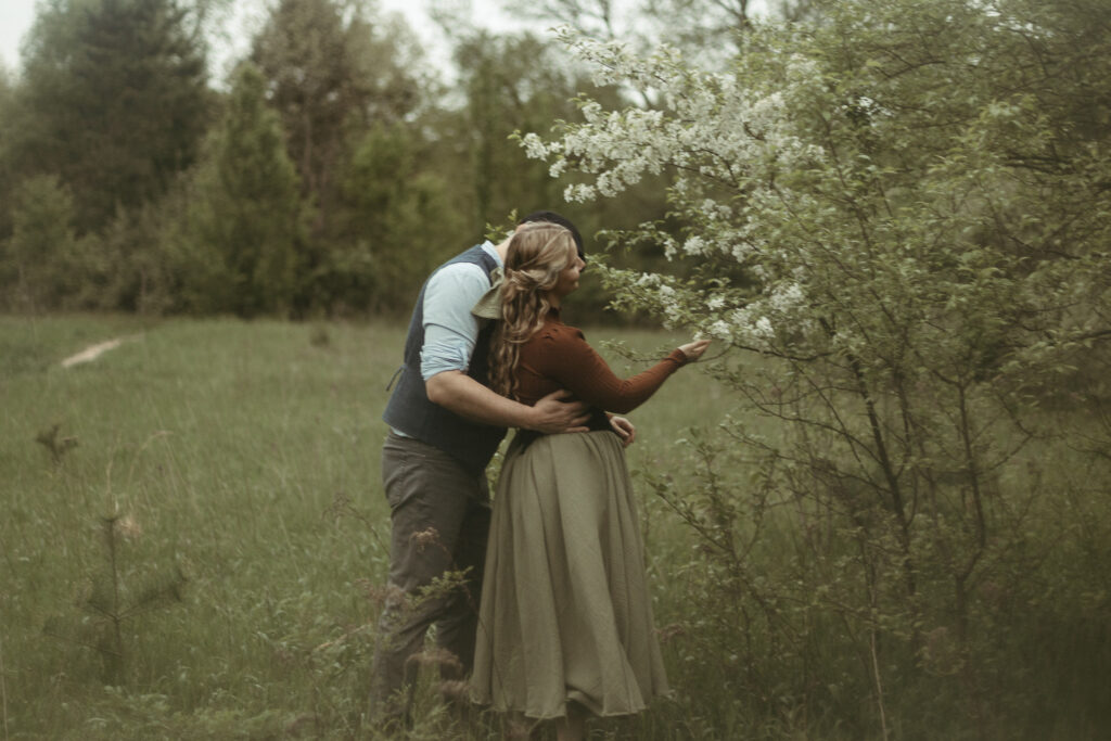 A woman skipping as she goes to a flower tree with her husband walking behind her at her engagement session in Michigan