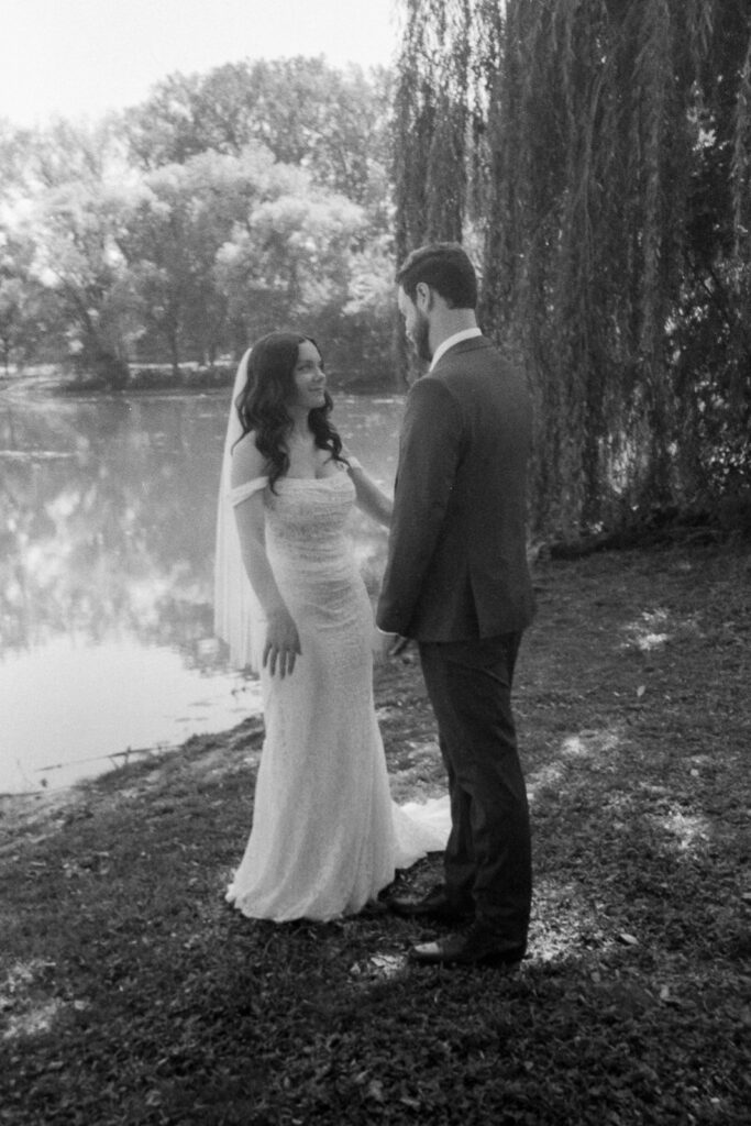 A bride and groom stand together beside a serene lake, surrounded by trees. The bride wears an elegant white gown, and the groom is in a suit. They look at each other lovingly under the soft, natural daylight.