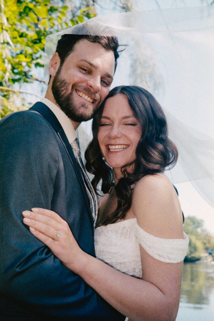 A newlywed couple smiles joyfully, embracing under a veil. The bride, in an off-shoulder dress, and groom, in a suit, are outdoors with greenery in the background. Both are laughing and looking content.