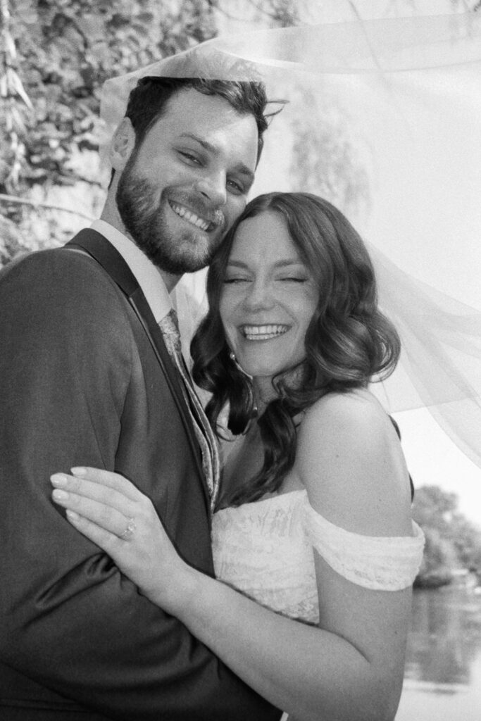 Happy couple embracing on their wedding day. The groom wears a dark suit, and the bride is in a lace off-shoulder gown with a veil. They are smiling joyfully outdoors, with trees and soft light in the background. The photo has a vintage black and white effect.