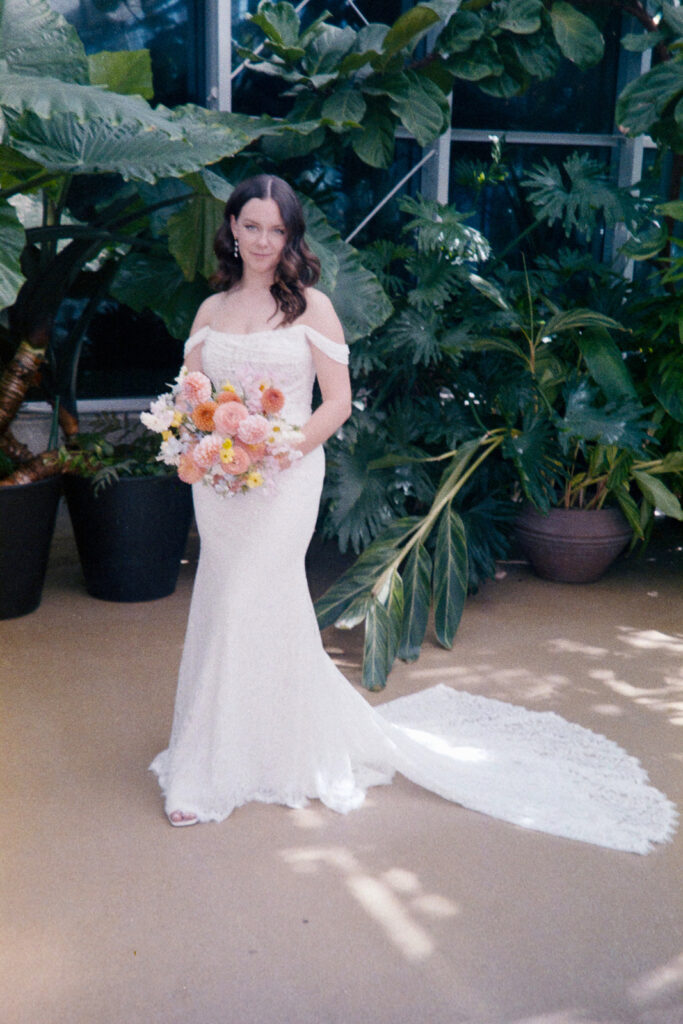 A woman in a long, off-shoulder white dress holds a colorful bouquet of flowers. She stands on a beige floor surrounded by lush green plants. The light casts soft shadows around her.