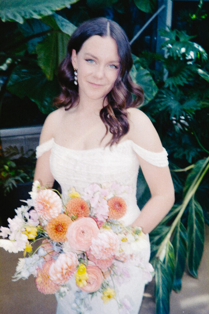 A woman in a white off-shoulder dress holds a colorful bouquet of flowers, including orange and pink blossoms. She stands in front of lush green foliage, with a gentle smile and long, wavy brown hair cascading down her shoulders.