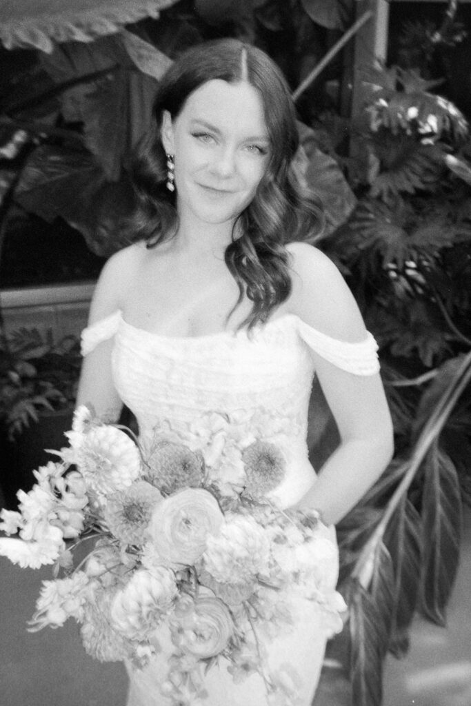 A woman in a white off-shoulder dress holds a bouquet of flowers, posing in front of lush greenery. The black-and-white image gives a classic and elegant feel, with her wavy hair and serene expression enhancing the graceful composition.