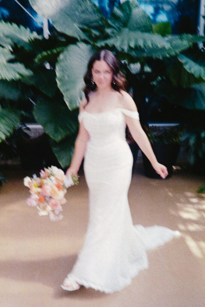 A woman in a white wedding dress holds a colorful bouquet. She is walking on a beige walkway surrounded by large green leaves, with dappled sunlight adding a dreamy effect.