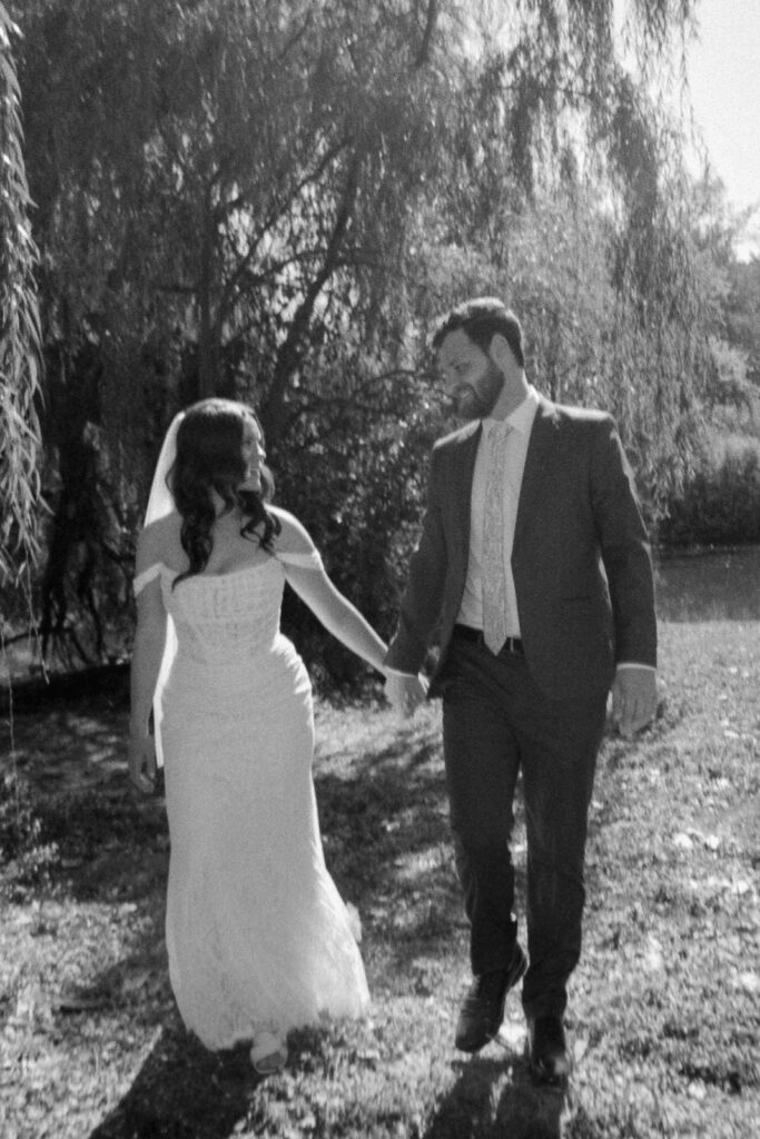 A black and white photo of a bride and groom walking hand in hand outdoors. The bride wears a long dress and veil, while the groom is in a suit. They are smiling at each other, with trees and a body of water in the background.