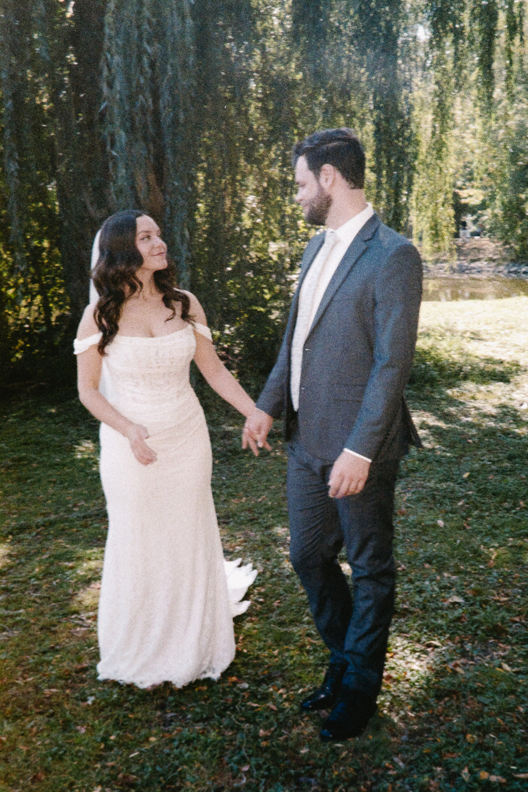 A bride in a white dress and a groom in a gray suit hold hands and gaze at each other while standing on a grassy area. The background features lush greenery and hanging willows, creating a serene and romantic atmosphere.