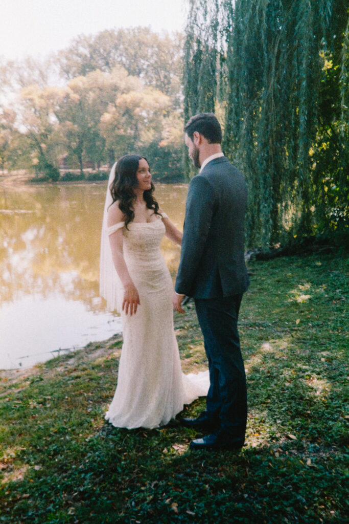 A bride and groom stand facing each other near a serene lake, surrounded by lush greenery and willow trees. The bride wears a white gown, and the groom is in a dark suit. Sunlight filters through the trees, creating a romantic atmosphere.