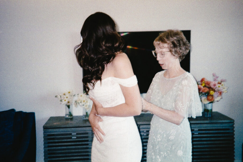 A bride with long hair stands in an off-shoulder white dress while an older woman, wearing glasses and a light blue dress, helps with the fitting. They are in a room with a dark piece of furniture and colorful flowers in vases.