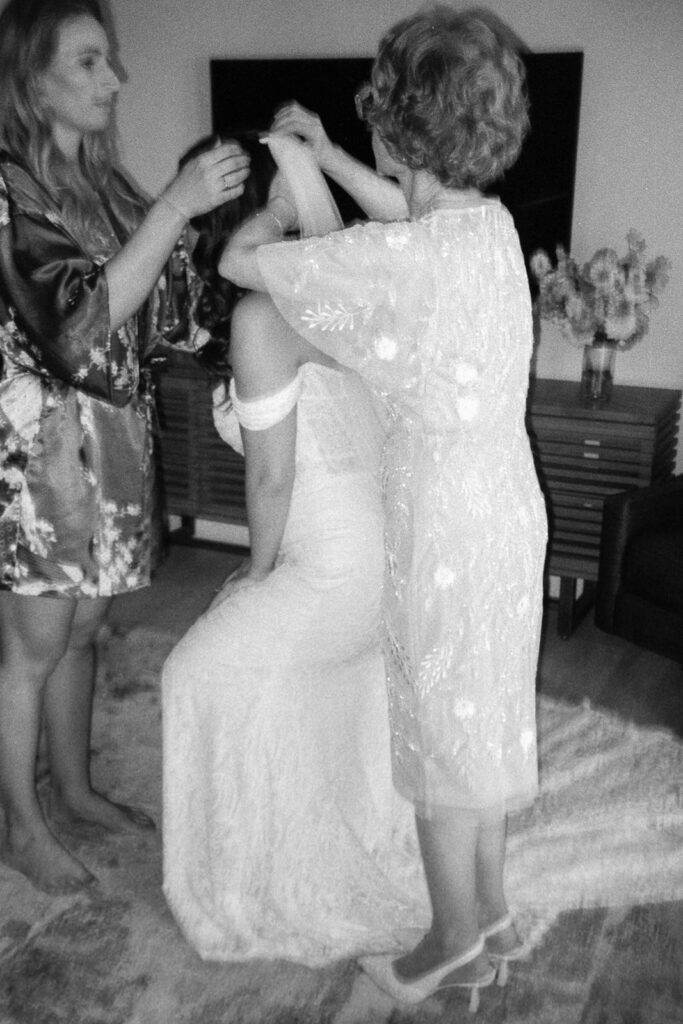 A black and white photo of a bride in a lace dress sitting on a chair, while two women adjust her veil. One woman is in a floral robe, and the other in a sheer, embroidered dress. Theyre in a living room with a TV and flowers in the background.