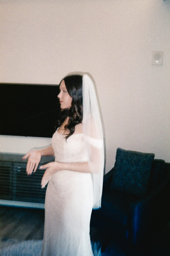 A bride in a white dress and veil stands in a room with a couch and a mounted TV. She has dark hair and appears to be gesturing with her hands. The room has neutral colors and a modern style.