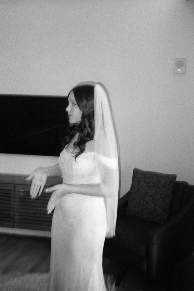 Black and white image of a woman in a wedding dress and veil standing indoors. She appears to be gesturing with her hands. There is a TV mounted on the wall and an armchair nearby.