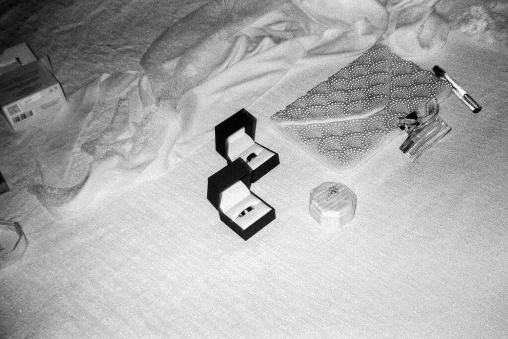 Black and white image of a table with an open ring box, a textured clutch bag, a pen, small perfume bottles, and a closed rectangular box. The table is covered with a cloth.