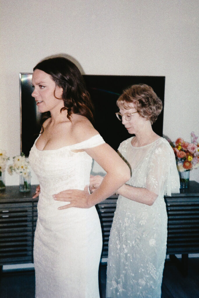 A bride in an off-shoulder wedding dress is being assisted by an older woman in a light-colored, floral-patterned dress. They are in a room with a flat-screen TV and a table decorated with flowers in the background.