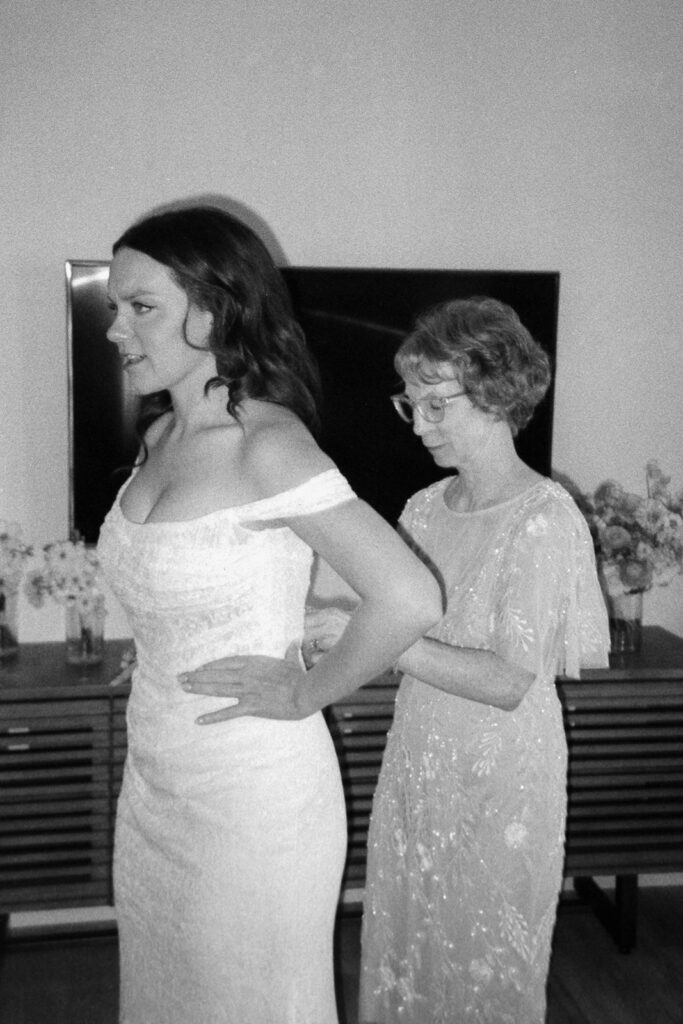 A black and white photo of a bride in an off-shoulder wedding gown being assisted by an older woman in a patterned dress. They stand in front of a TV, with floral arrangements on a wooden console behind them.