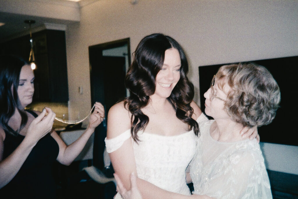 A bride in a white dress smiles while embracing an older woman in a sheer outfit. Another woman stands nearby, holding a necklace. The scene is set in a softly lit room.