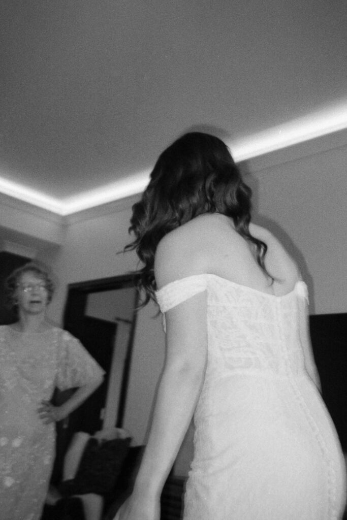 Black and white image of a woman in an off-shoulder dress facing away from the camera. Another woman, smiling, stands in the background. Theyre in a room with a bright ceiling light.