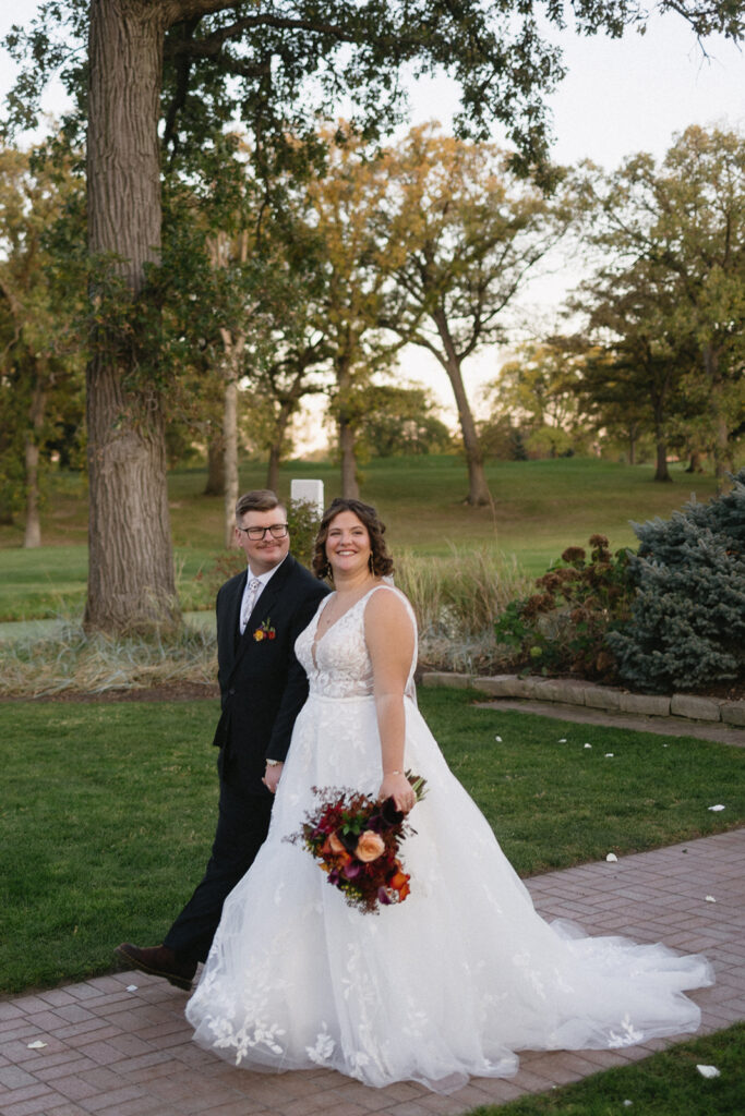 A couple walks hand in hand on a brick path through a park. The bride wears a white gown and holds a colorful bouquet, while the groom is in a black suit. Trees with autumn leaves surround them under a clear sky.