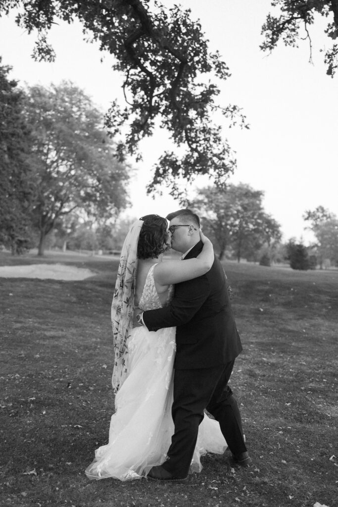 A couple in wedding attire embraces on a grassy field surrounded by trees. The bride wears a flowing gown with a long veil, while the groom is in a dark suit. They kiss under a trees branches in a serene outdoor setting.