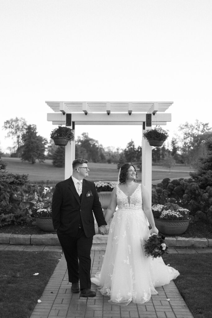 A couple holds hands while walking on a brick path under an archway. The bride wears a floral gown and holds a bouquet, and the groom is in a dark suit. The background shows greenery and potted plants. Its a black and white photo.