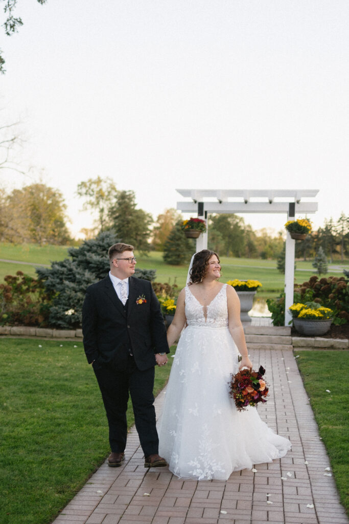 A couple in wedding attire walks hand in hand on a brick path outdoors. The bride wears a white lace gown with a bouquet of autumn flowers, and the groom wears a black suit with a patterned tie. Theres a white arbor and greenery in the background.