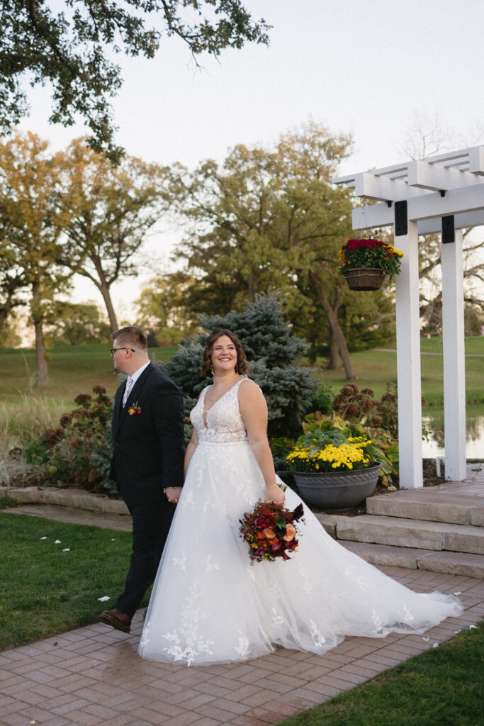 Bride in a white gown holds a bouquet while standing on a brick path next to a groom in a dark suit. They stand near a white pergola with hanging plants in a garden setting, surrounded by greenery and trees.