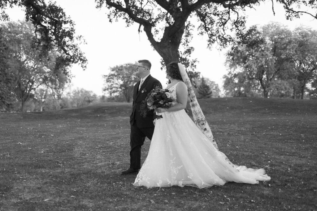 A bride and groom stand together in a grassy park, with the bride holding a bouquet and wearing a long, flowing dress. The groom is in a suit. Both are gazing into the distance, with a large tree and scattered leaves in the background. The image is in black and white.