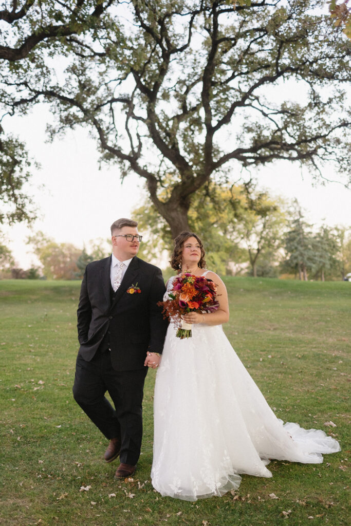 A bride and groom walk hand in hand on a green lawn. The bride wears a white gown and holds a bouquet of red and orange flowers. The groom is in a black suit with a boutonniere. A large tree stands in the background.