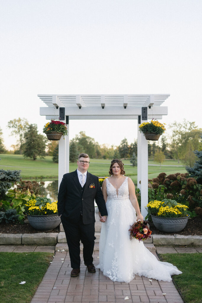 A couple stands under a white pergola in a garden setting. The person on the left wears a black suit, and the person on the right is in a white lace dress holding a bouquet. Flower pots with yellow blooms decorate the scene.