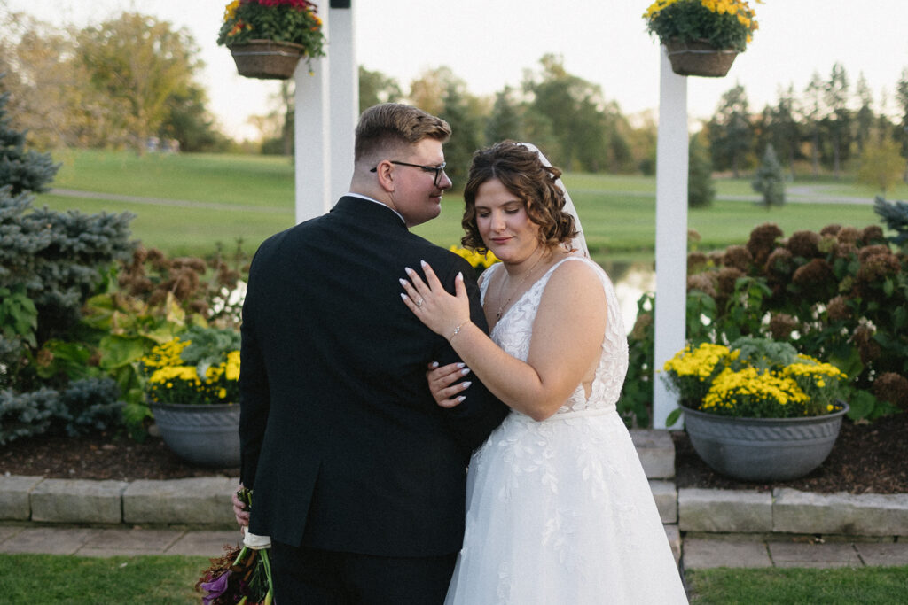 A bride and groom embrace outdoors in a garden setting. The bride is in a white gown, and the groom is in a black suit. They stand in front of a trellis with hanging plants, surrounded by greenery and flowers, with a pond and trees in the background.