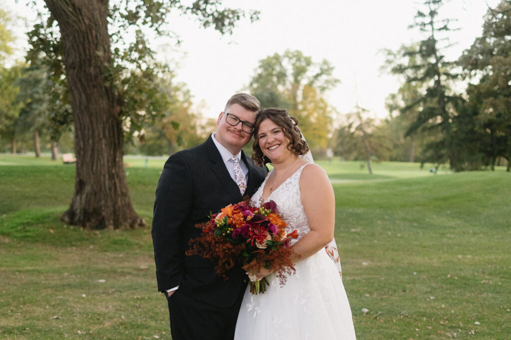 A happy couple in wedding attire pose outdoors on a grassy field. The bride holds a vibrant bouquet of flowers. Trees and greenery are visible in the background, creating a serene, natural setting.