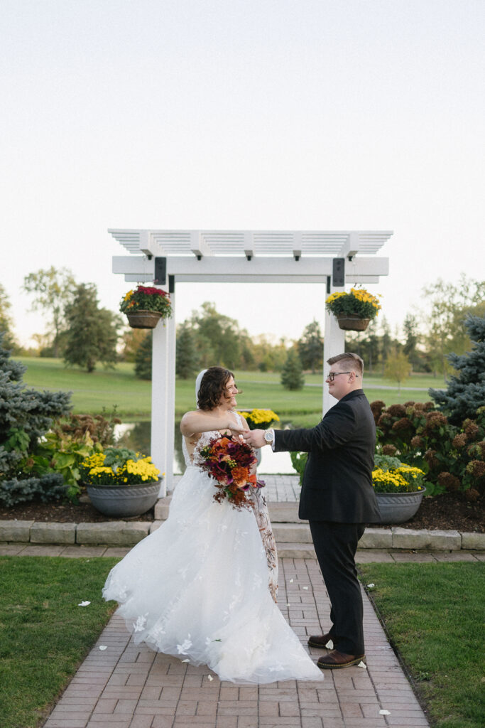 A bride in a white gown and a groom in a black suit share a dance outdoors under a white pergola. The bride holds a bouquet of red and yellow flowers. They stand on a brick path with greenery and yellow flowers around.