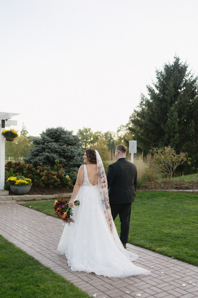 A bride in a white gown with a long veil and a groom in a dark suit walk hand in hand on a brick path. The bride holds a bouquet of flowers. They are outdoors in a garden setting with trees and greenery in the background.