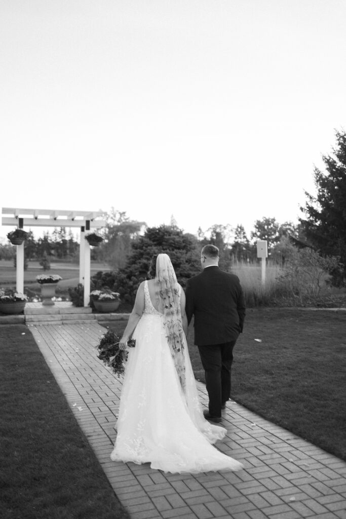 A bride in a flowing gown and veil walks hand in hand with a groom in a suit along a brick path. They are outdoors, with greenery and trees around, near a pergola. The scene is serene, captured in black and white.