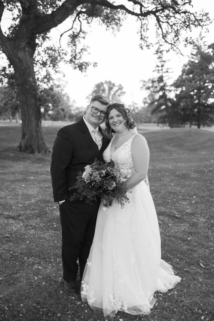 A black-and-white photo of a smiling couple on their wedding day. The groom is in a suit, and the bride is in a white gown holding a bouquet. They stand closely under a tree, with a grassy landscape in the background.