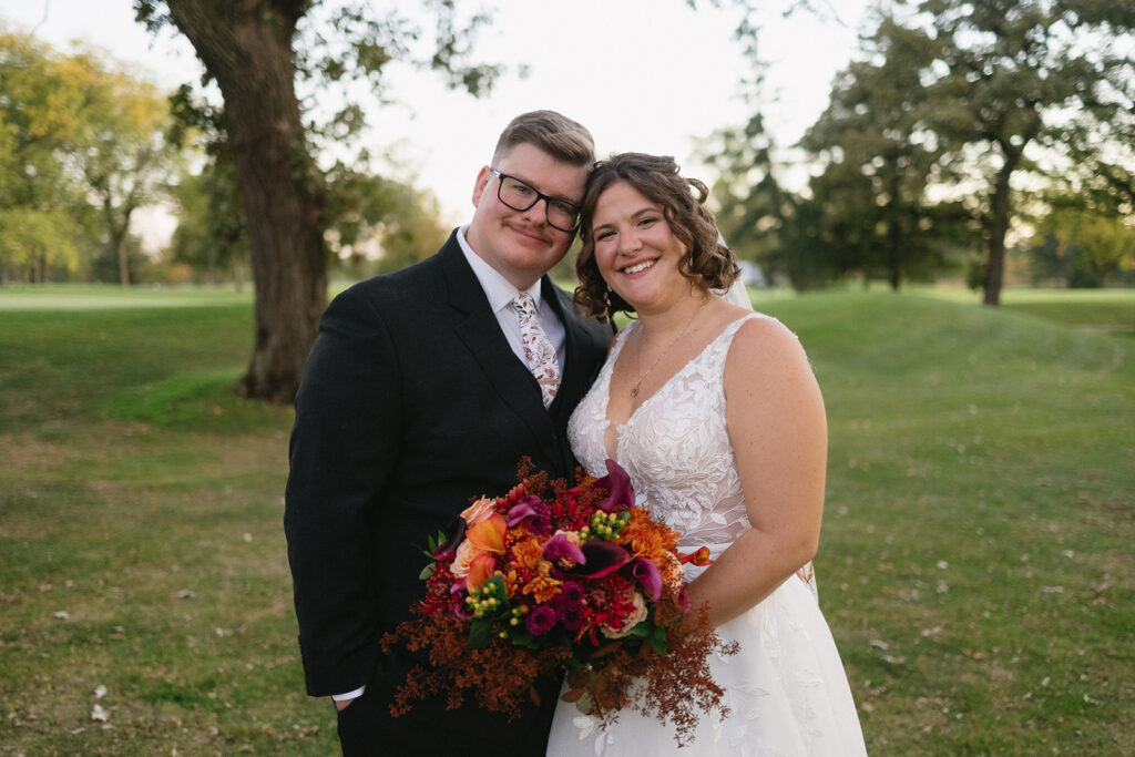 A bride and groom stand close together in a grassy outdoor setting. The bride, in a white gown, holds a colorful bouquet. Both are smiling warmly at the camera, with trees and a soft, natural background behind them.