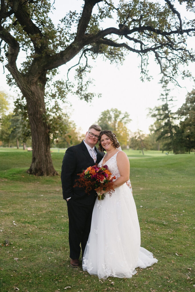 A bride and groom smile under a large tree in a park. The bride holds a bouquet of red and orange flowers, wearing a white gown, while the groom is in a black suit. The background features grass, trees, and a clear sky.