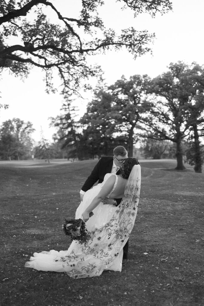 A black-and-white photo of a newlywed couple sharing a romantic kiss in a park. The bride holds a bouquet, and her long veil is flowing. Theyre surrounded by large trees under a clear sky. The groom is gently dipping the bride.