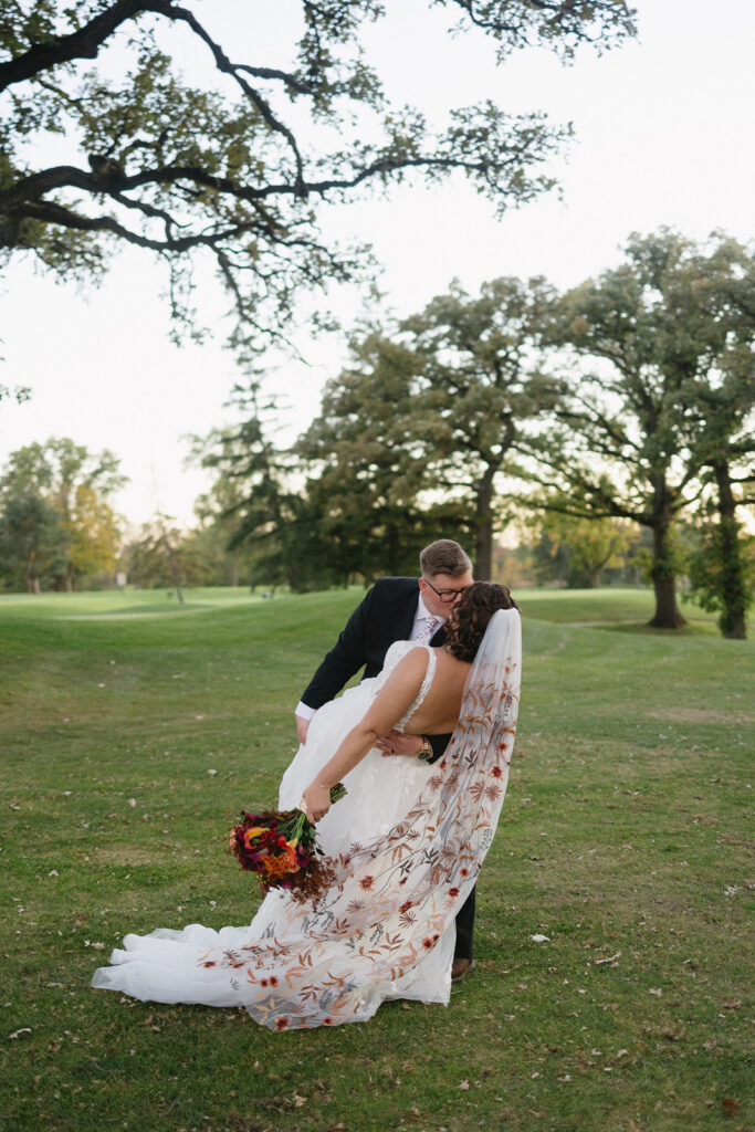 A newlywed couple shares a romantic dip on a grassy field surrounded by trees. The bride wears a white dress with floral embroidery and holds a bouquet of red and orange flowers. The groom is in a black suit and leans in for a kiss.