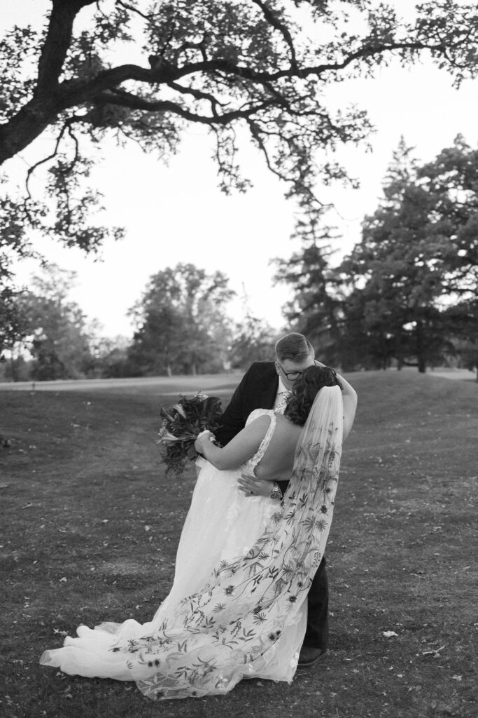 A couple embraces under a large tree on a grassy field. The bride wears a long, floral-patterned veil and holds a bouquet, while the groom is in a dark suit. Trees and open sky form the backdrop. The image is in black and white.