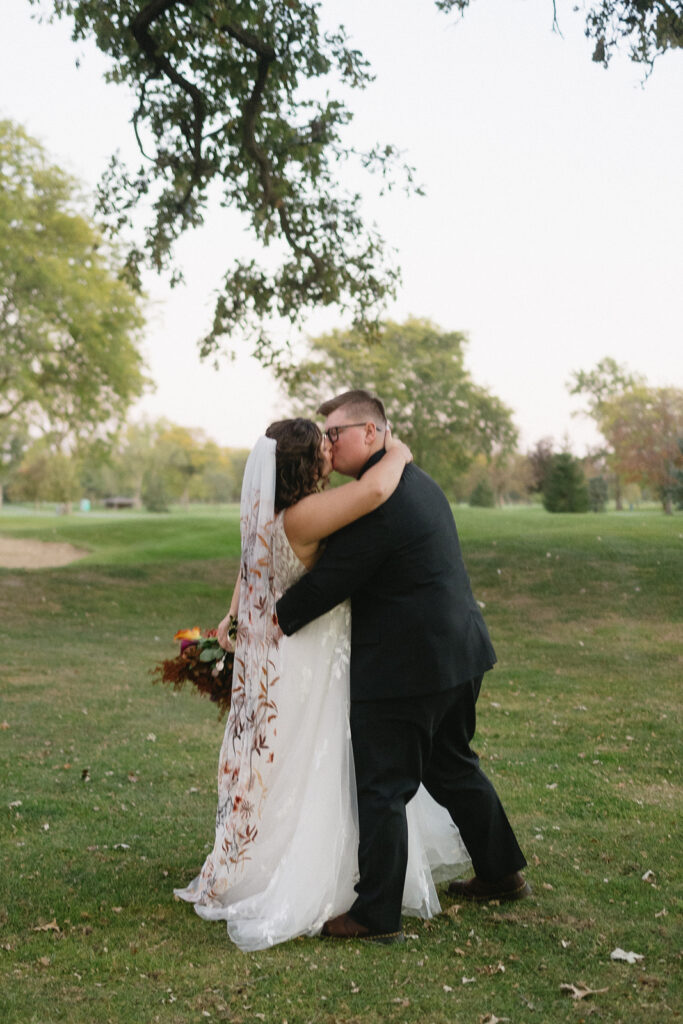 A bride and groom share a kiss outdoors on a grassy lawn. The bride is in a white dress holding a bouquet with red and yellow flowers. The groom is in a black suit. Trees with green foliage are in the background.