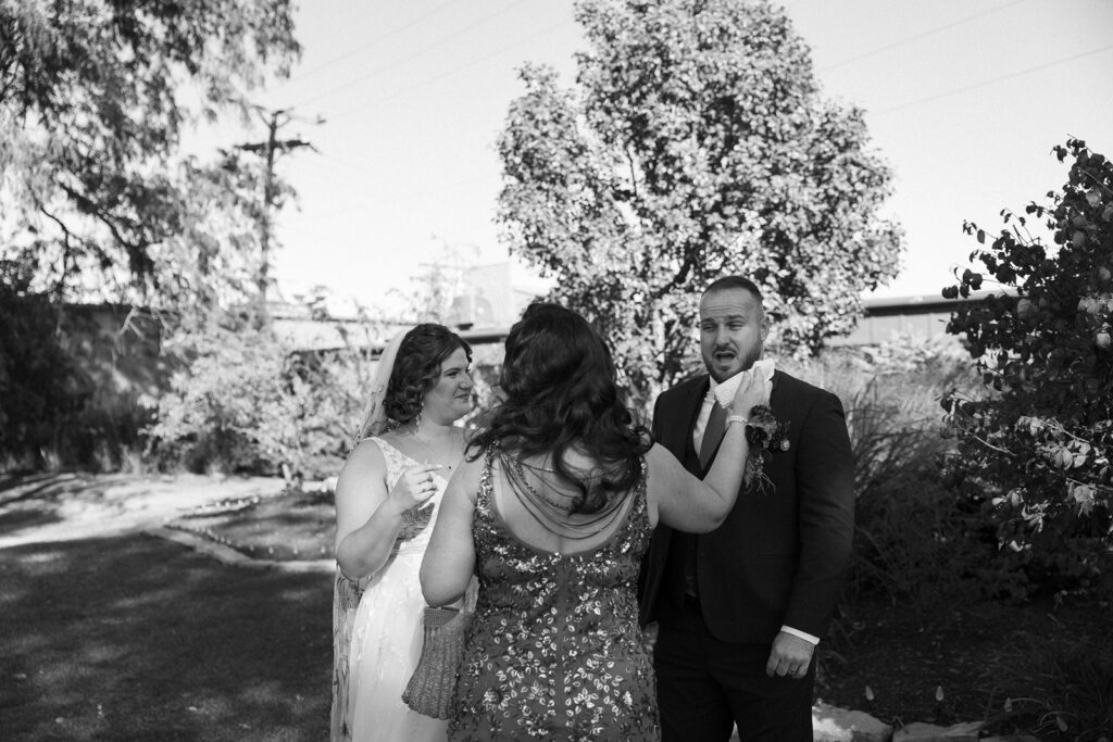 A black and white photo of three people outdoors, with trees and plants in the background. A man in a suit and two women in dresses are talking and appear engaged in a lively conversation.