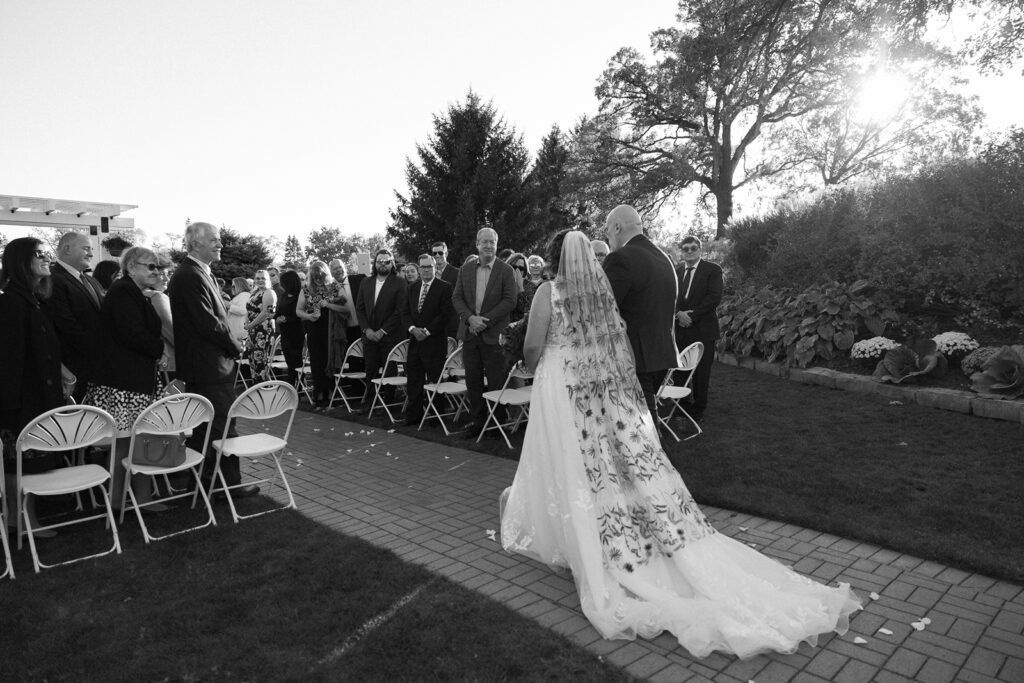 A bride and groom walk down an outdoor aisle lined with chairs and guests on either side. The bride wears a long, detailed dress, and the groom is in a suit. Sunlight filters through trees in the background. The scene is in black and white.