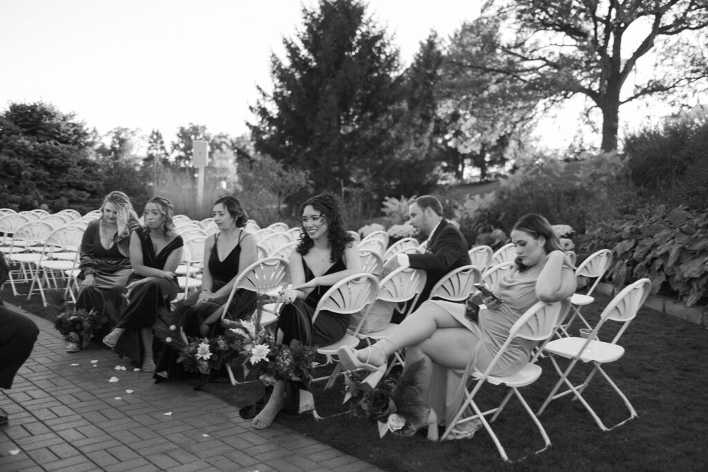 A black and white photo of six people sitting on outdoor folding chairs at an event. They are dressed formally, with flower arrangements visible near the chairs. The background includes trees and plants, giving a serene, garden-like atmosphere.