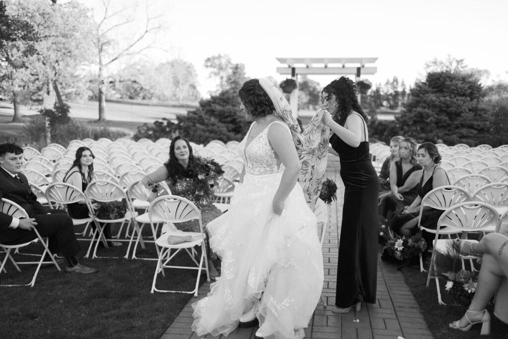 A bride in a wedding gown stands in the middle of an outdoor venue with white chairs, holding hands with another woman in a dark dress. Guests are seated nearby. The setting is surrounded by trees under a clear sky.