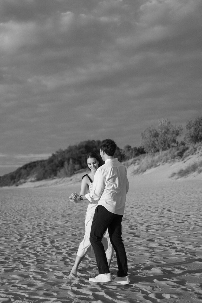 A couple dances barefoot on a sandy beach, under a cloudy sky. The woman wears a light dress, and the man is in a shirt and pants. They appear joyful and carefree, with a dune and trees in the background. The image is in black and white.