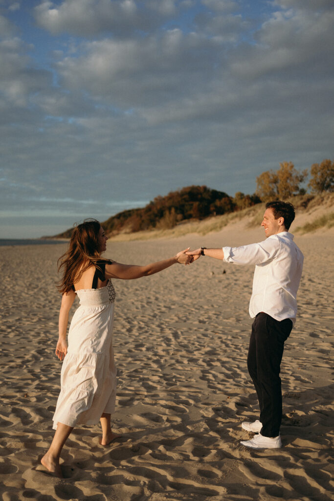 A couple joyfully dances on a sandy beach under a partly cloudy sky. The woman, in a white dress, and the man, in a white shirt and dark pants, hold hands. Trees and dunes are visible in the background.