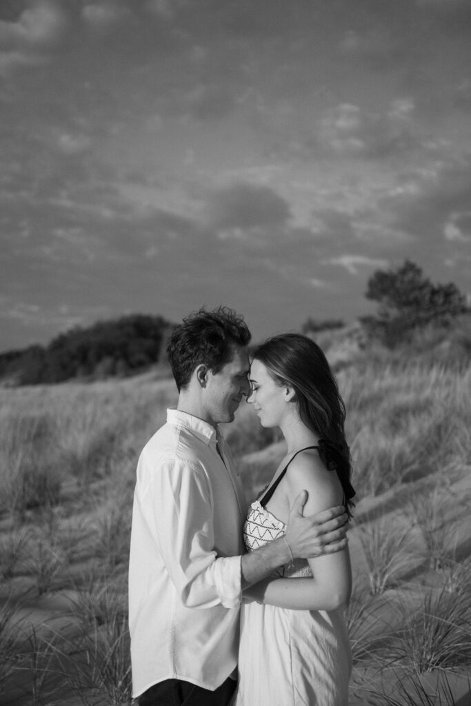 A black and white photo of a couple embracing on a sandy beach with grassy dunes. They are facing each other closely, exuding a sense of intimacy. The sky is partly cloudy, creating a soft, romantic atmosphere.