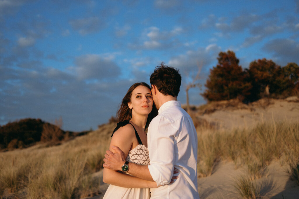 A couple stands in a sandy, grassy area under a blue sky with scattered clouds. The woman faces the camera, wearing a patterned dress, while the man, in a white shirt, has his back to the camera, embracing the woman. Trees are visible in the background.