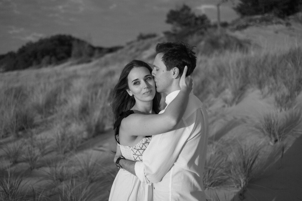 A couple embraces on a sandy beach. The woman with long hair looks at the camera while the man kisses her cheek, his eyes closed. They wear light-colored clothing, and dunes with grass are in the background. The image is in black and white.